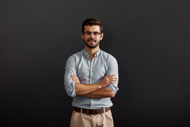 Project manager fiducioso e giovane uomo barbuto che guarda la telecamera con un sorriso e tiene le braccia