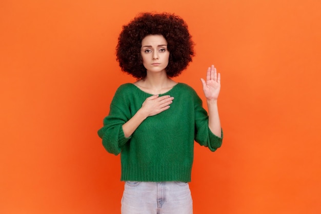 Prometto donna seria con acconciatura afro che indossa un maglione verde in piedi alzando la mano e dicendo giuramento fedeltà giuramento giuramento fedeltà Indoor studio girato isolato su sfondo arancione