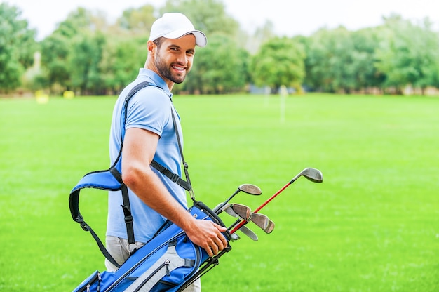 Pronto a giocare. Vista posteriore del giovane giocatore di golf felice che porta la sacca da golf con i conducenti e si guarda alle spalle mentre si trova in piedi sul campo da golf