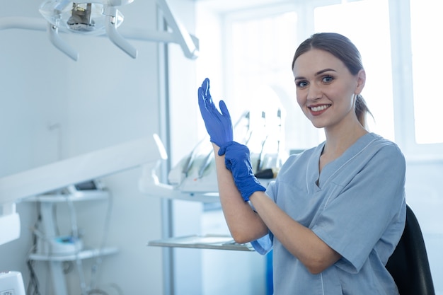 Pronto a lavorare. Bella giovane femmina dentista che indossa guanti di gomma e sorride alla telecamera mentre si prepara per il trattamento