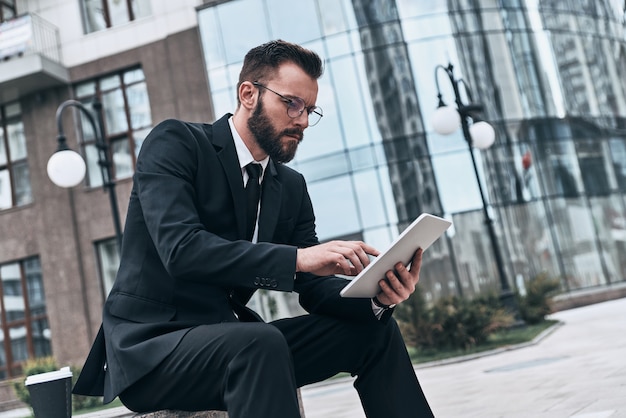 Pronto a lavorare sodo. Giovane di bell'aspetto in abito completo che utilizza tablet digitale mentre è seduto all'aperto