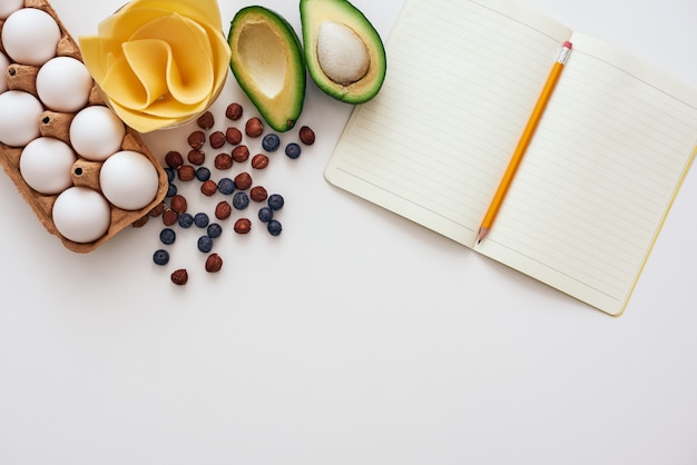 Pronto a scrivere una ricetta. Il libro di cucina e le uova sono distesi sul tavolo vicino a bacche essiccate, formaggio e avocado