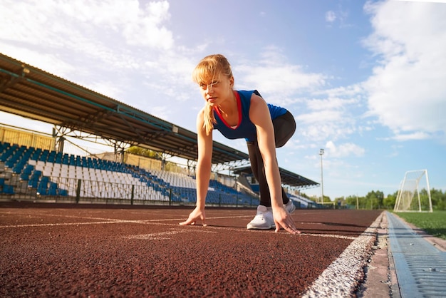 Pronto per partire Atleta bionda femminile in una maglietta blu e leggings sulla linea di partenza di una pista dello stadio che si prepara per una corsa La ragazza di forma fisica si prepara a correre da un inizio basso