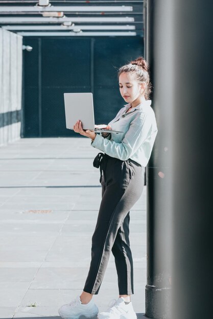 Proprietario della donna di affari che tiene computer portatile vicino al deposito dell'ufficio, all'aperto. Viaggiare, lavorare all'aperto in città, stare in strada fuori sotto il sole, lavorare al computer portatile, felice e guardare la telecamera