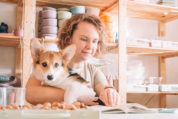 Proprietario di donna d'affari allegra dai capelli ricci con cane corgi seduto rilassante e leggendo un libro sul posto di lavoro al chiuso Piatti in silicone per piccoli affari per bambini Negozio per ufficio confortevole a casa