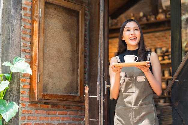 Proprietario femminile del caffè sorridente con felicità positiva pronta ad aprire per la donna asiatica cliente con il vassoio del regalo della mano del grembiule e la tazza di caffè caldo benvenuto condident in piedi all'ingresso del coffeeshop