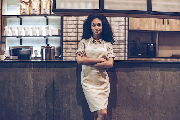 Proprietario sicuro del caffè. Giovane donna africana allegra in grembiule che tiene le braccia incrociate e guarda la telecamera con un sorriso mentre si trova al bancone del bar bar