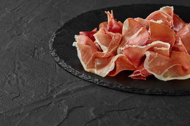 Prosciutto gourmet affettato manualmente su tavola di ardesia di pietra nera su sfondo grigio scuro Colpo del primo piano Vista laterale Copia spazio