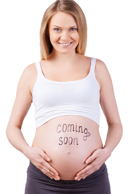 Prossimamente. Immagine ritagliata della donna incinta con presto segno sul suo ventre in piedi isolato su bianco