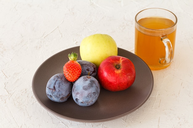 Prugne, mele, una fragola sul piatto e una tazza di tè sulla superficie bianca strutturata