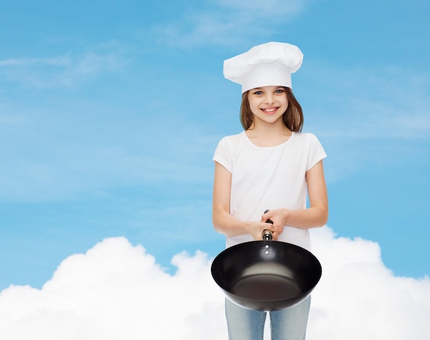 pubblicità, infanzia, cucina e persone - ragazza sorridente in t-shirt bianca e cappello da cucina che tiene padella su sfondo blu cielo