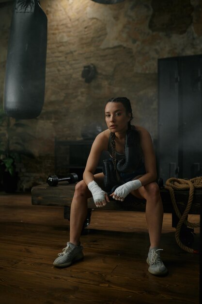 Pugile femminile in bendaggio da boxe che porta guanti sulle spalle seduto sulla panchina