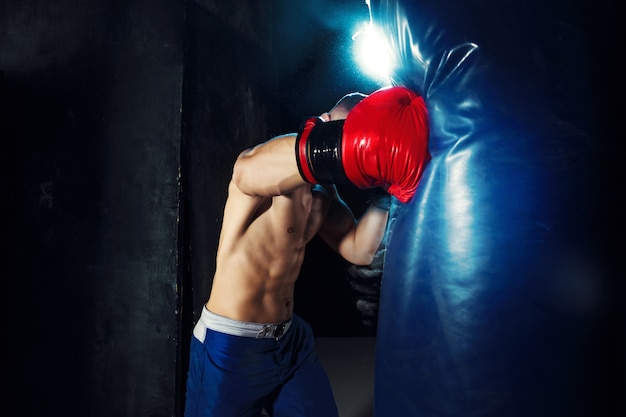 Pugile maschio dell'atleta che perfora un sacco da boxe con illuminazione tagliente drammatica in uno studio scuro