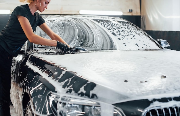 Pulisce il veicolo che è in sapone bianco La moderna automobile nera viene pulita da una donna all'interno della stazione di lavaggio auto