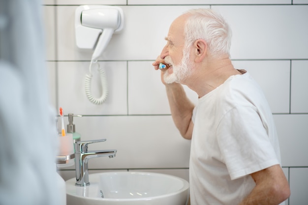 Pulizia dei denti. Uomo dai capelli grigi in maglietta bianca che pulisce i suoi denti