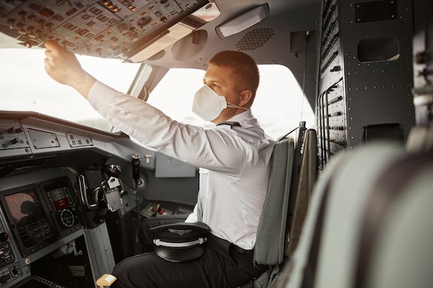 Pulsante di tornitura pilota maschio sul jet dell'aeroplano passeggeri. Interno della cabina di pilotaggio in aereo con cruscotto e navigazione aerea. L'uomo europeo indossa l'uniforme e la maschera medica. Aviazione civile. Concetto di viaggio aereo