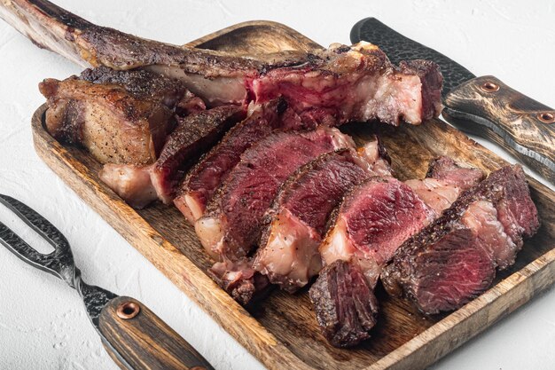 Punta di costola tomahawk alla brace con sale al rosmarino ed erbe aromatiche mediamente cotta su tavola da portata in legno su sfondo di pietra bianca