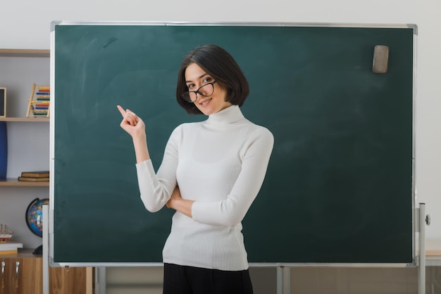 punti felici a lato giovane insegnante femminile che indossa occhiali in piedi davanti alla lavagna in classe