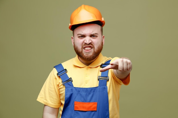 Punti tesi alla fotocamera giovane costruttore uomo in uniforme isolato su sfondo verde