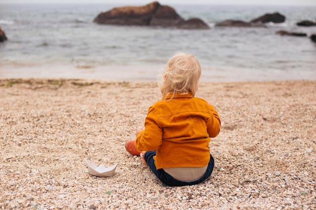 Punto di vista posteriore del ragazzo che gioca con la barchetta di carta che si siede sulla spiaggia vicino al mare in autunno o in estate