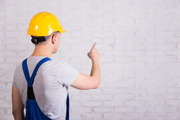 Punto di vista posteriore dell'uomo in uniforme blu del costruttore che punta al muro di mattoni bianco