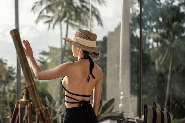 Punto di vista posteriore della donna in vestito nero che sta vicino al telescopio su un balcone che guarda la vista sull'oceano