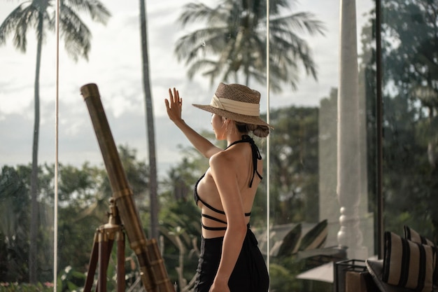 Punto di vista posteriore della donna in vestito nero che sta vicino al telescopio su un balcone che guarda la vista sull'oceano.