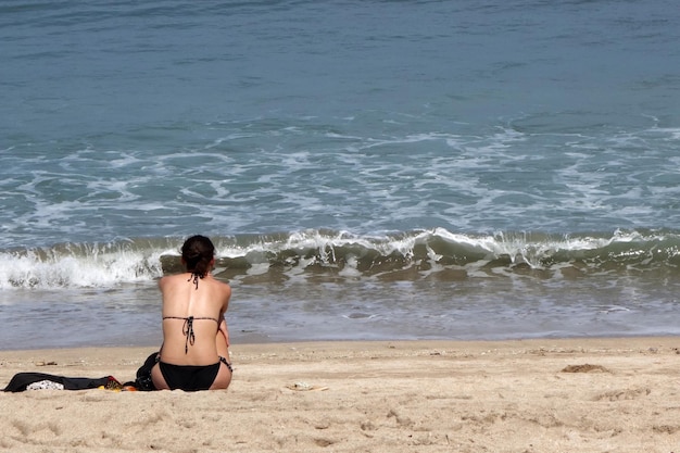 Punto di vista posteriore della donna sexy in bikini che si siede sulla spiaggia tropicale di sabbia bianca