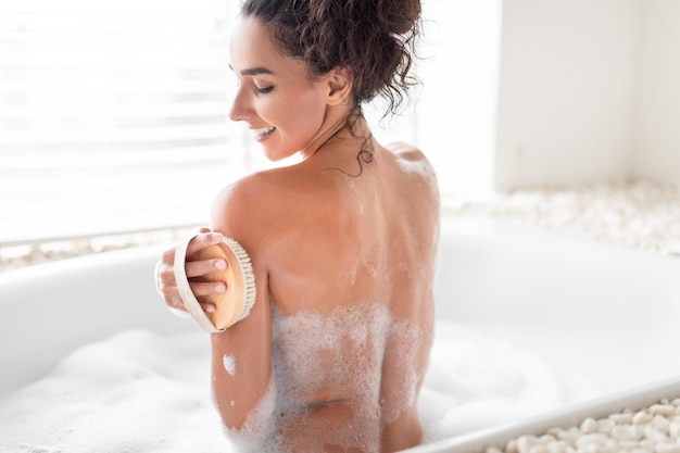 Punto di vista posteriore della giovane donna adorabile che fa il massaggio linfatico della spazzola asciutta che sfrega la sua pelle nel bagno frizzante