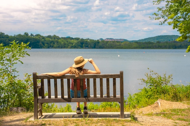 Punto di vista posteriore della giovane donna attraente che si rilassa su una panchina all'aperto davanti ad un lago