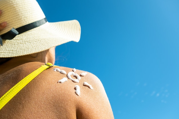 Punto di vista posteriore della giovane donna che si abbronza sulla spiaggia con la crema della protezione solare a forma di sole sulla sua spalla. Protezione dai raggi UV e protezione solare