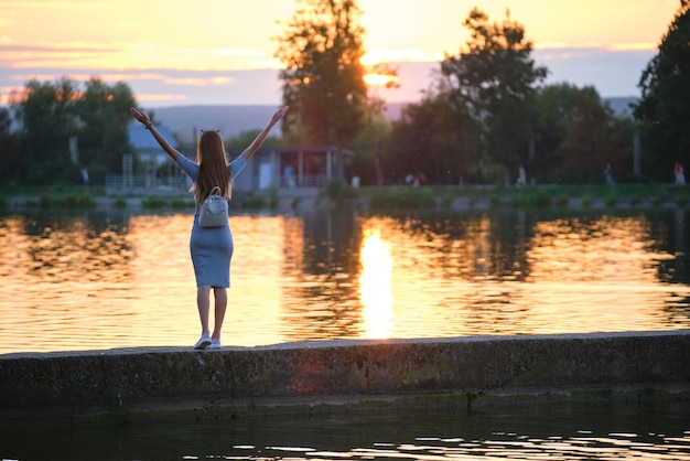 Punto di vista posteriore della giovane donna sola che sta da sola sulla riva del lago alzando le mani in alto godendosi la calda serata. Concetto di successo e meditazione.