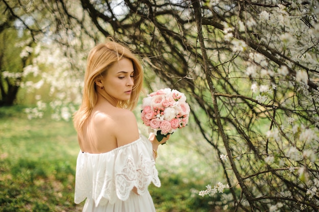 Punto di vista posteriore di giovane donna bionda in un vestito bianco con il mazzo vicino al ciliegio di fioritura