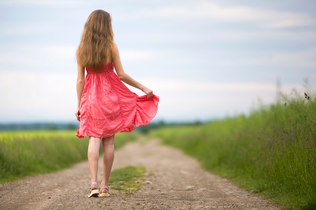 Punto di vista posteriore di giovane donna esile romantica in vestito rosso con capelli lunghi che cammina dalla strada a terra lungo il campo verde il giorno di estate soleggiato sullo spazio della copia del cielo blu.