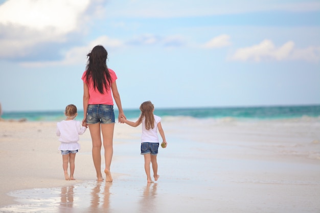 Punto di vista posteriore di giovane madre e delle sue figlie sveglie che camminano alla spiaggia tropicale