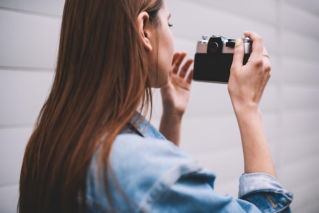 Punto di vista posteriore di giovane ragazza alla moda dei pantaloni a vita bassa dei capelli del brunette che fa le immagini tramite la retro macchina fotografica vicino al muro