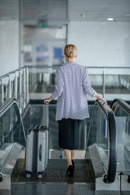 Punto di vista posteriore di un passeggero femminile alla moda con la valigia del carrello che guida la scala mobile all'aeroporto