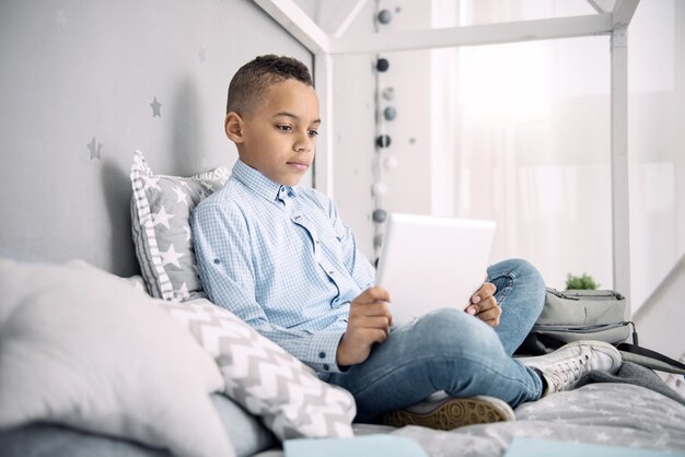 Punto interessante. Focalizzato ragazzo afroamericano utilizzando tablet mentre è seduto sul letto