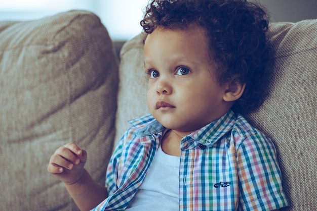 Puoi annegare nei suoi occhi. Ritratto di piccolo bambino africano che guarda lontano mentre è seduto sul divano di casa