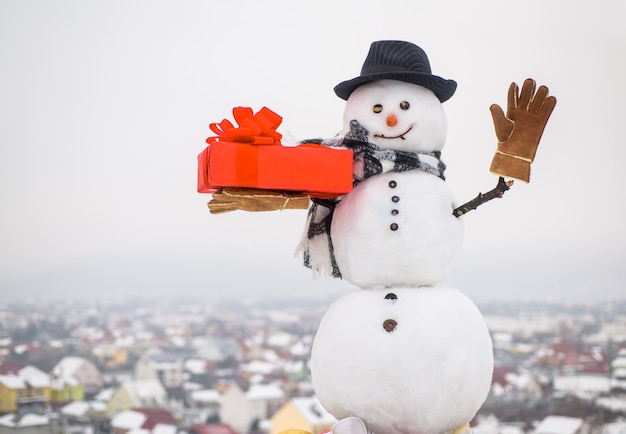 Pupazzo di neve felice con regalo agitando la mano pupazzo di neve con scatola regalo pupazzo di neve con cappello sciarpa guanti natale