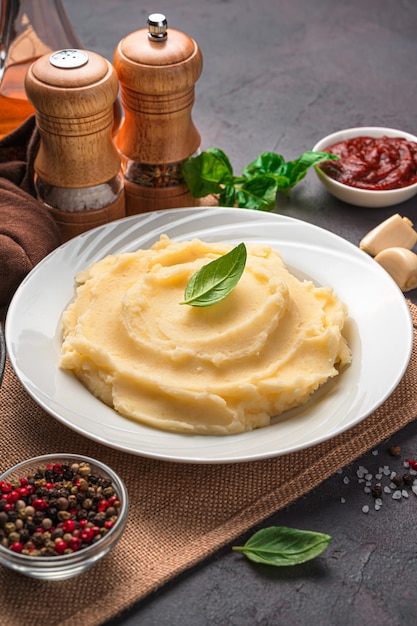 Purè di patate in un piatto bianco su uno sfondo di spezie, salsa e basilico. Vista laterale, verticale.