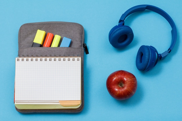 Quaderno aperto su astuccio borsa con pennarelli colorati e pennarello, cuffie e mela su sfondo blu. Vista dall'alto. Torna al concetto di scuola. Materiale scolastico