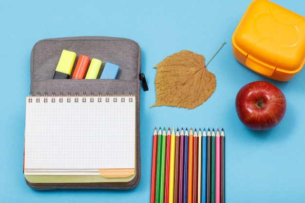 Quaderno aperto su astuccio portamatite con pennarelli colorati e pennarello, scatola per il pranzo, mela, foglia secca e matite colorate su sfondo blu. Vista dall'alto. Torna al concetto di scuola. Materiale scolastico