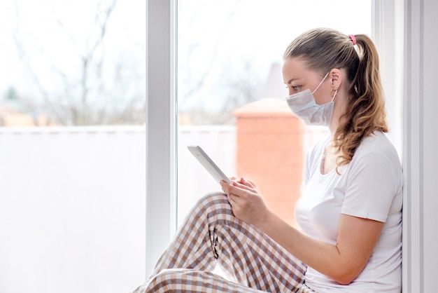 Quarantena a casa. Donna caucasica in congedo per malattia in maschera medica con tablet nelle sue mani. Ordina la consegna di cibo e medicine.