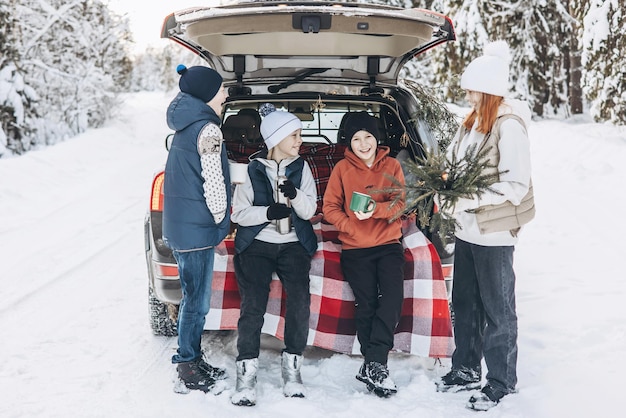 Quattro amici adolescenti ragazzi e ragazze con thermos seduti nel bagagliaio dell'auto decorato per Natale e Capodanno Picnic invernale nella foresta innevata Viaggio su strada e viaggi locali Amici uscire insieme