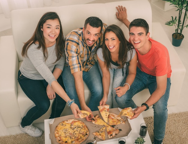 Quattro amici allegri che bazzicano in un appartamento. Mangiano la pizza e guardano la fotocamera.