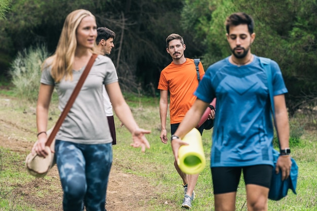 Quattro amici che parlano rilassati mentre trasportano materassini e borse da yoga passeggiando nella foresta