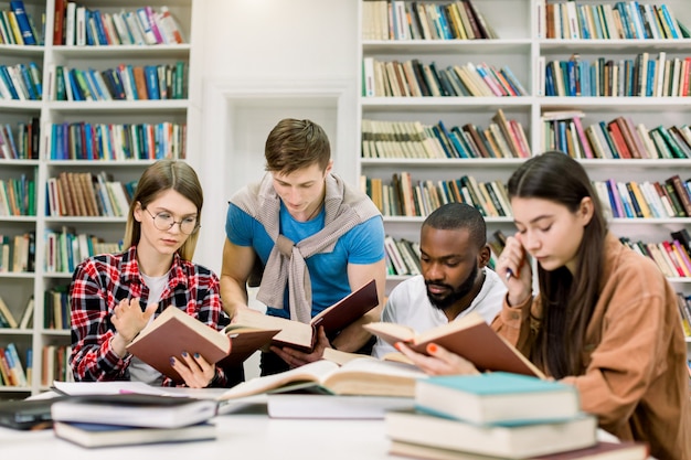 Quattro amici multietnici intelligenti concentrati seduti al tavolo della biblioteca universitaria e leggendo libri insieme, preparandosi per l'esame o la lezione, facendo compiti a casa