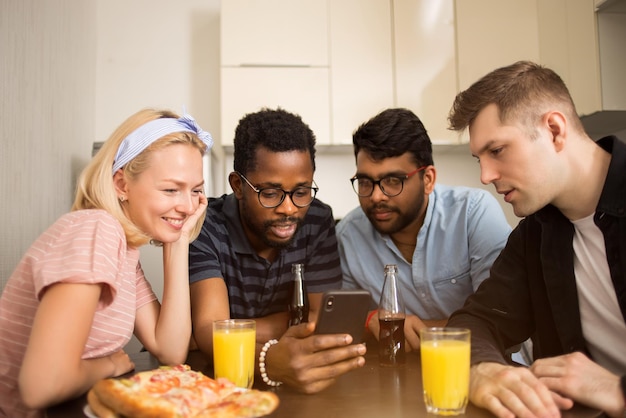 Quattro amici seduti a tavola, mangiano pizza, bevono bibite, leggono messaggi di testo su smartphone, testano insieme nuove applicazioni telefoniche. Studenti multiculturali che si divertono nella cucina dell'ostello.