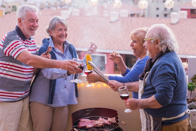 quattro anziani attivi in pensione si divertono in terrazza a casa cucinando qualche barbecue tutti sorridono e stanno insieme in amicizia sotto una bella giornata di sole. vacanza e relazione persone concetto people